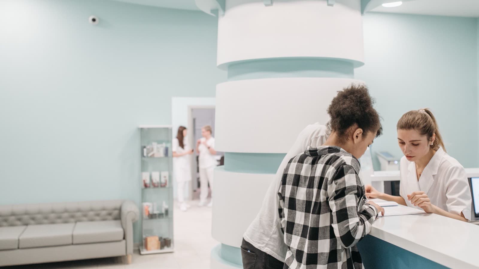 Two visitors at the reception desk of a medical centre, with a waiting area behind them