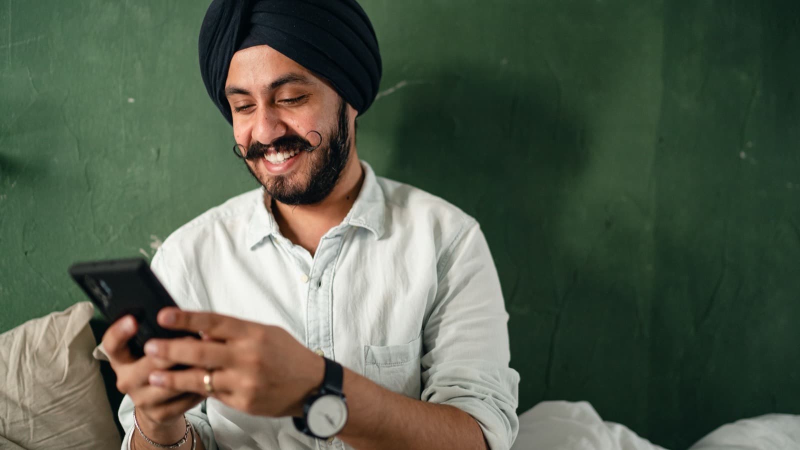 A smiling man sitting on a bed, typing on his phone